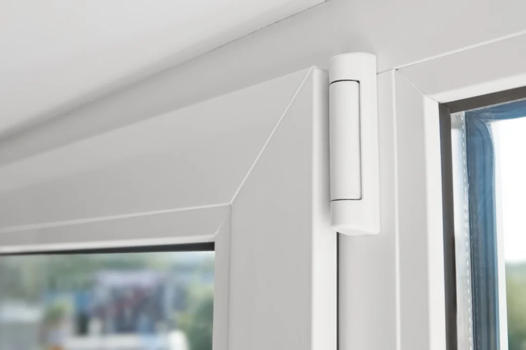 White vinyl window hinge joins a closed sash to its frame, securing double-glazed glass in a bright interior corner with a blurred outdoor scene visible beyond the pane.