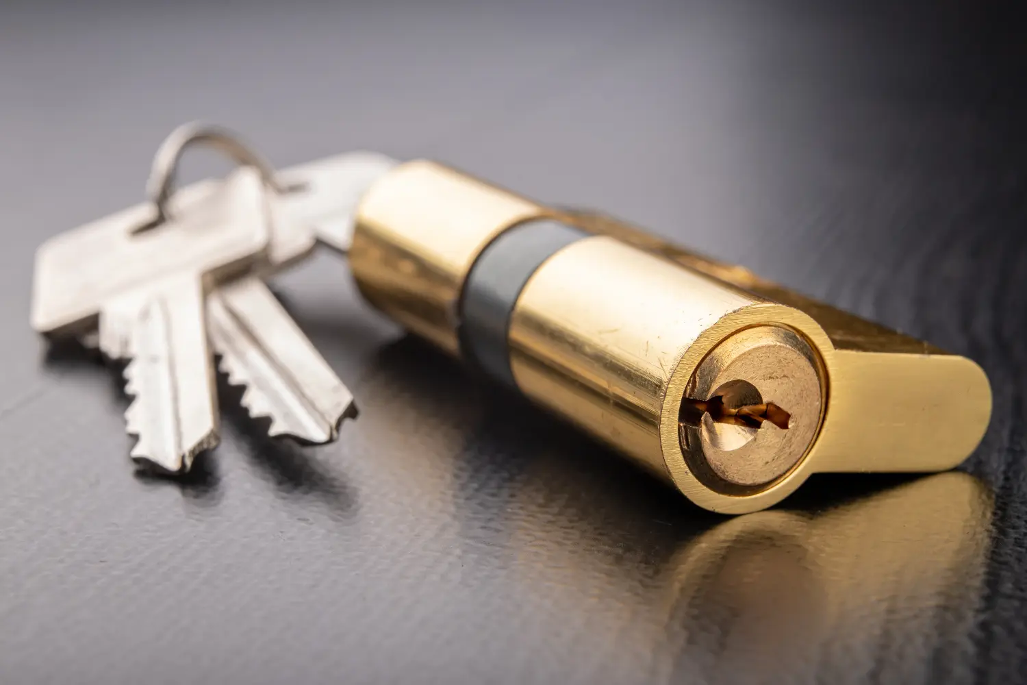 A brass cylinder lock rests beside two silver keys on a dark textured surface, reflecting light and revealing the lock's keyway.