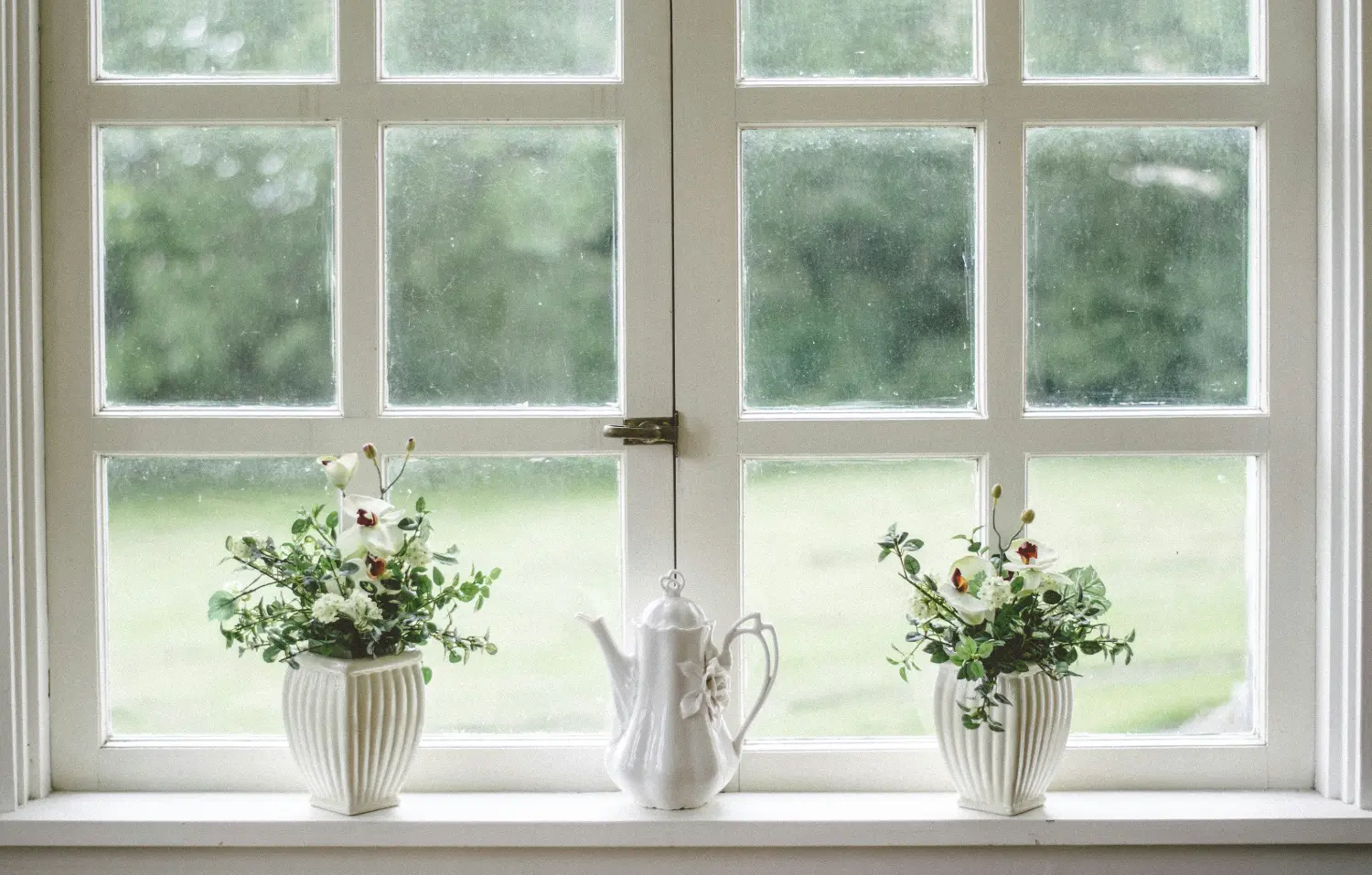 White ceramic teapot centered between two potted flower arrangements, resting on a windowsill, framed by a multi-pane window with a softly blurred green garden beyond.