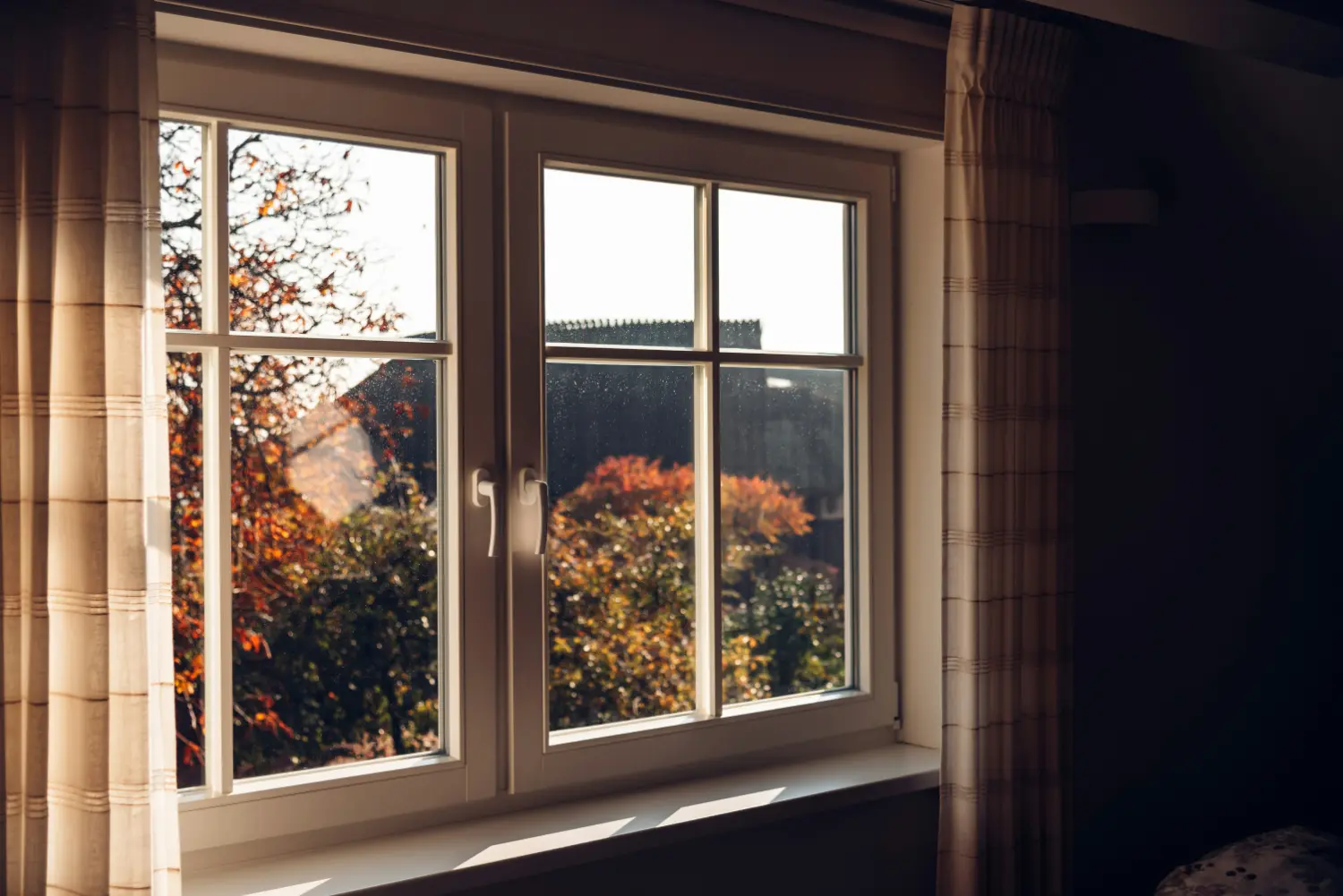 A white-framed casement window frames sunlit autumn foliage and a dark rooftop outside, casting warm light onto a simple windowsill inside, flanked by beige striped curtains in a quiet room.