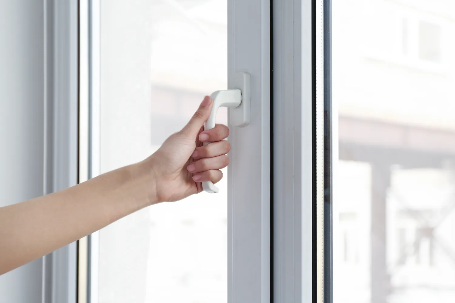 A hand grips a white window handle, turning it to open a closed double-glazed frame against a bright, blurred exterior seen through clear glass.