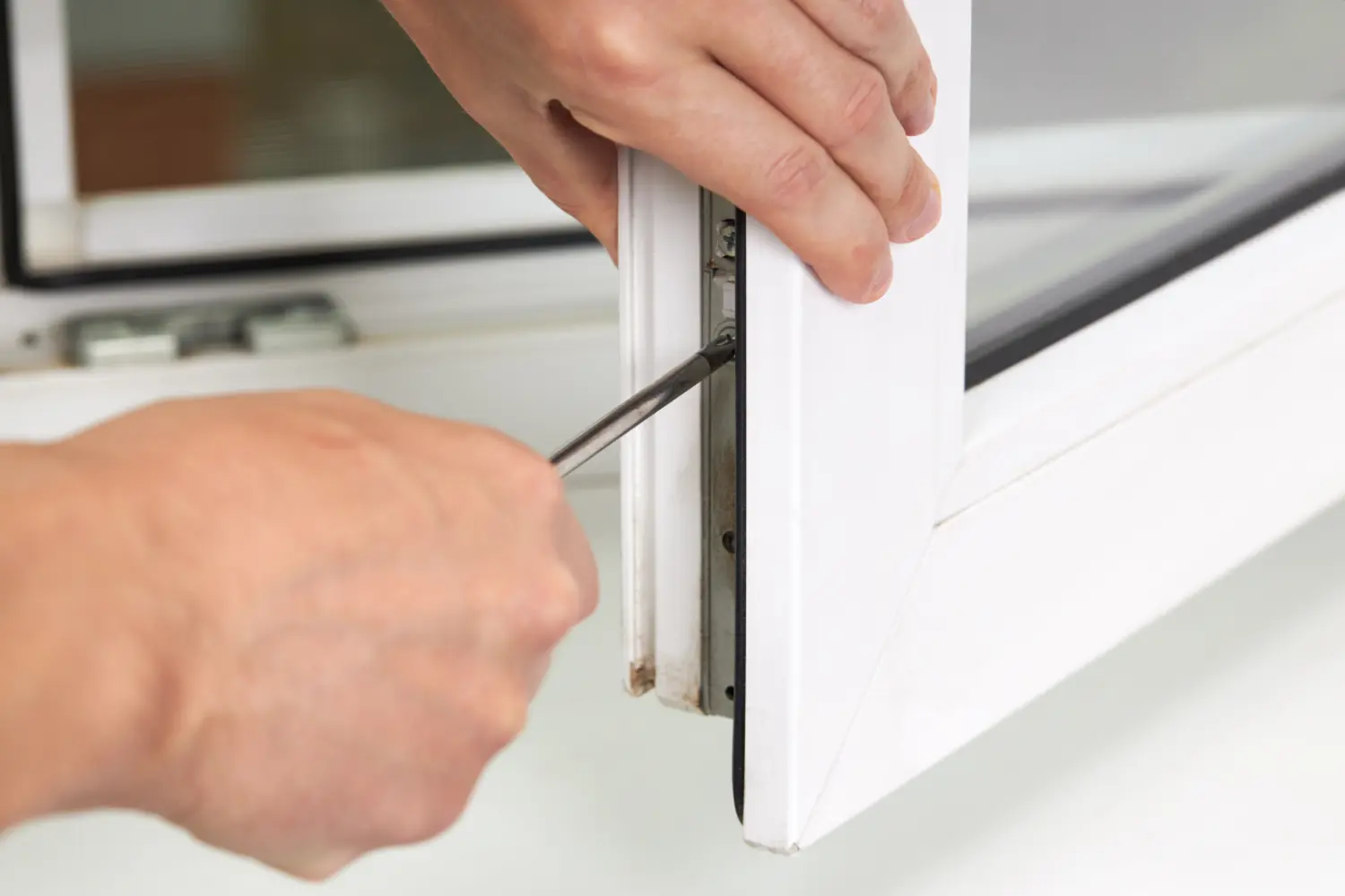 PVC window hinge being adjusted with a screwdriver by two hands while the open white window sash sits above a light indoor windowsill.